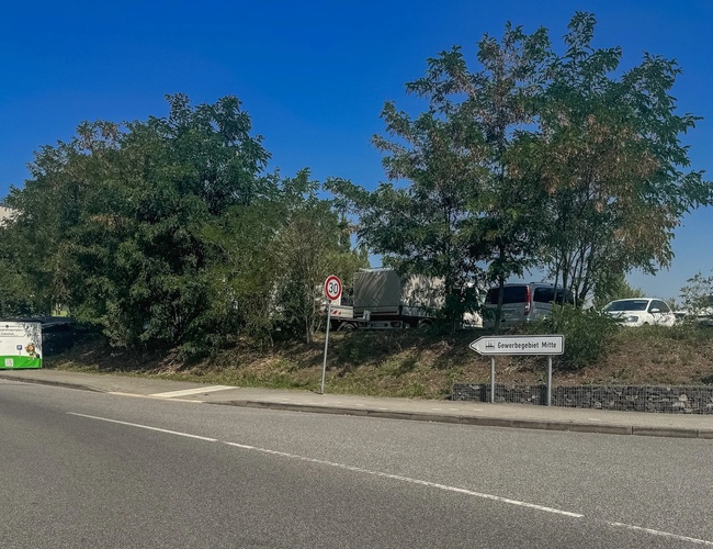 Parking Mein-Flughafenparkplatz-Frankfurt P1. Widok z ulicy na ogrodzony teren parkingu i baner reklamowy obok drogi.