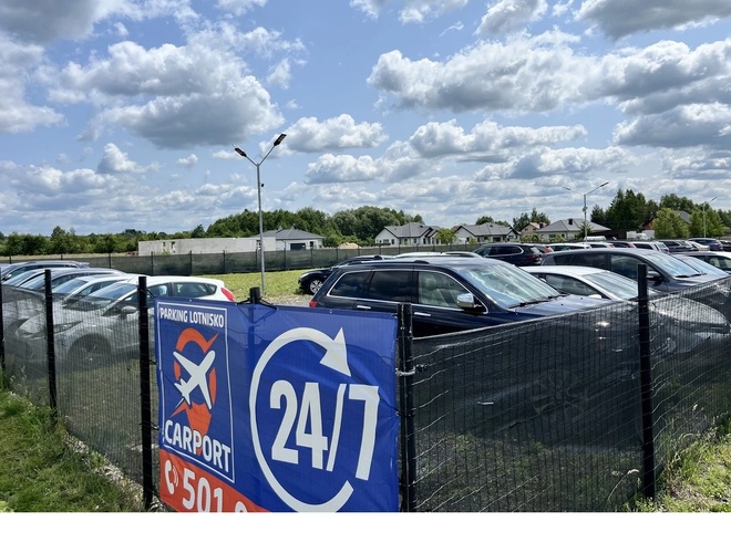 Parking CARPORT RDO - Ogrodzony teren z samochodami osobowymi. Widoczny baner z logo i napisem 24/7. Latarnie stoją wśród zaparkowanych aut.