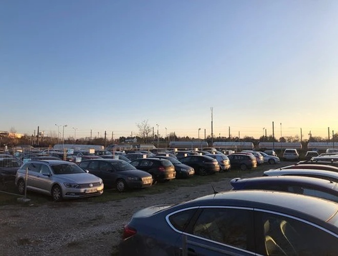 Parktiger Airport Parking 4 - Na parkingu stoją samochody na trawiastej nawierzchni. Teren jest ogrodzony i dobrze oświetlony. W tle widać tory kolejowe.