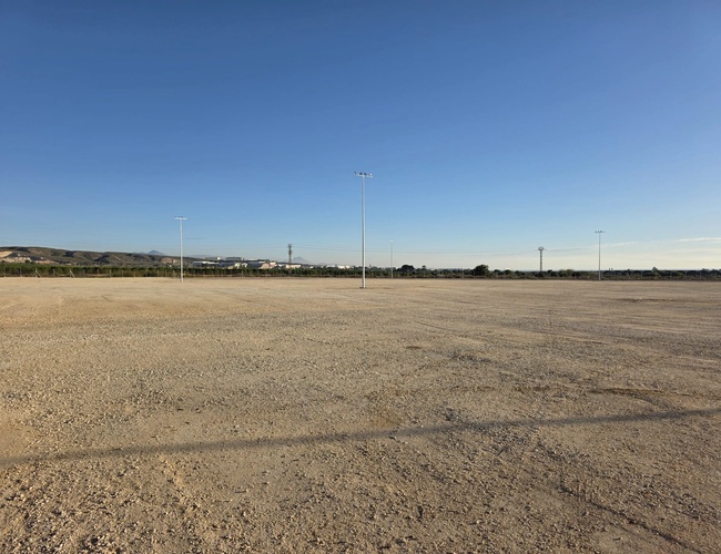 Rozległy, pusty żwirowy teren pod jasnym, błękitnym niebem, widok na horyzont z niskimi zabudowaniami.