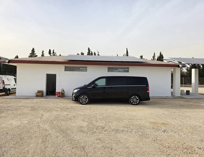 Biały budynek z jednym czarnym samochodem zaparkowanym na żwirowym podjeździe, w tle drzewa i niebo z delikatnymi chmurami.