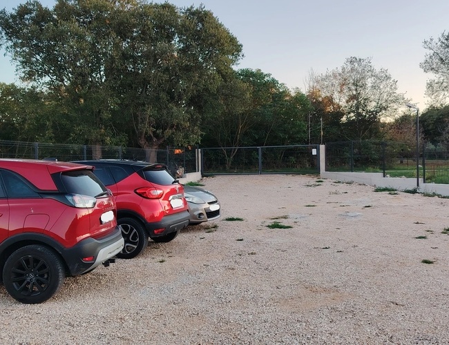 Samochody zaparkowane na parkingu ze żwirowym podłożem. W tle widoczne ogrodzenie oraz drzewa. Na końcu parkingu brama wjazdowa.