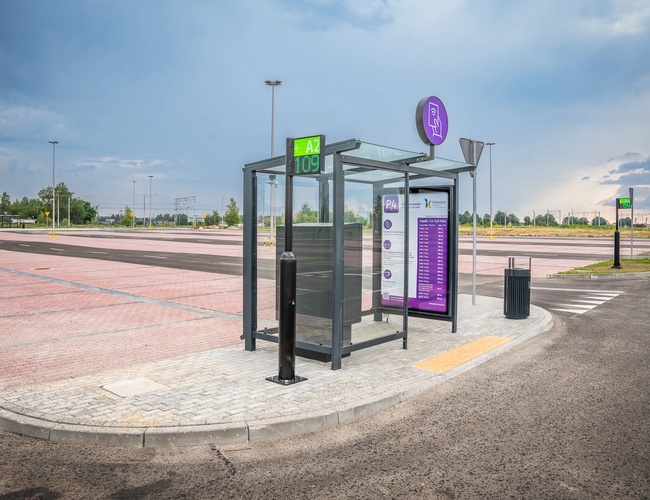 Parkometr z wiatą, tablicą informacyjną i koszem na śmieci na tle dużego, pustego parkingu.
