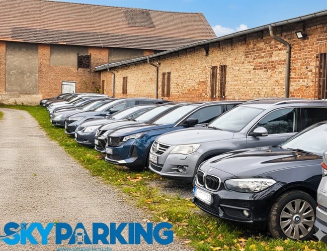 Rzędy samochodów zaparkowanych wzdłuż wąskiego, utwardzonego przejazdu między starymi murowanymi budynkami, w słoneczny dzień.