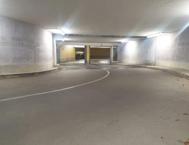 Kręta droga w podziemnym parkingu, oświetlona sztucznym światłem, z widocznym wyjściem na końcu tunelu.