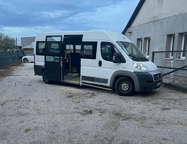 Biały bus zaparkowany na żwirowym placu obok budynku, drzwi pojazdu są otwarte