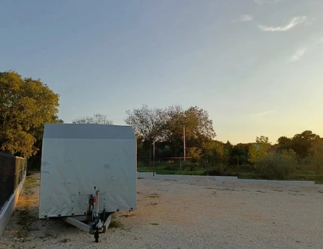 Niewielka przyczepa lub kontener ustawiony na pustym, żwirowym terenie otoczonym drzewami o zachodzie słońca