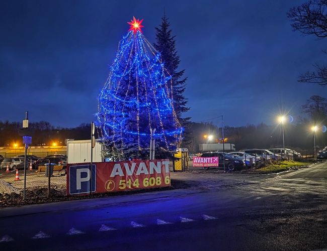 Widok oświetlonego parkingu nocą, wjazd na parking. Na terenie parkingu znajduje się ogromna choinka ozdobiona lampkami.