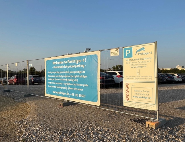 Niebieski znak umieszczony na ogrodzeniu w parkingu z informacją o parkingu oraz instrukcją jego użytkowania.