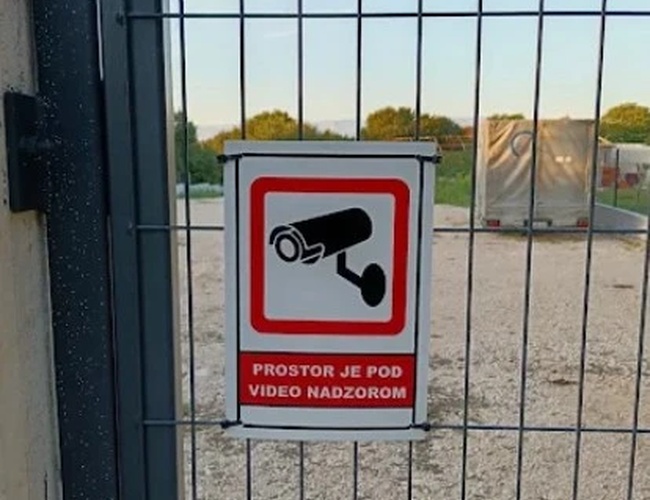 Brama z metalową siatką, na której widoczny jest znak informujący o monitoringu na parkingu.