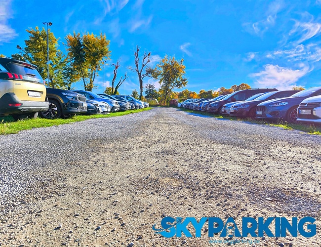 Ujęcie wykonane z niskiej perspektywy, przedstawiające szutrową nawierzchnię i rzędy samochodów zaparkowanych na otwartym terenie przy słonecznej pogodzie.