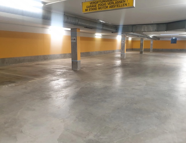 Pusta przestrzeń w podziemnym parkingu z betonową podłogą i żółtymi oznaczeniami na ścianach.