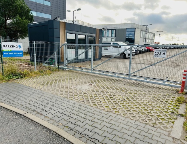 Parking Piątka - Parking z ogrodzeniem i stojącymi autami. W tle nowoczesne budynki.
