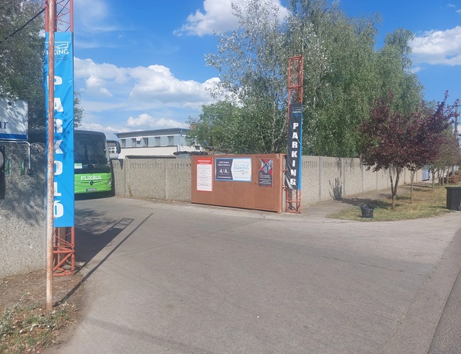 Parking BUDCAR - wjazd na parking, widoczna szeroka brama. Po bokach banery z nazwą parkingu. Dookoła ogrodzenie oraz drzewa.