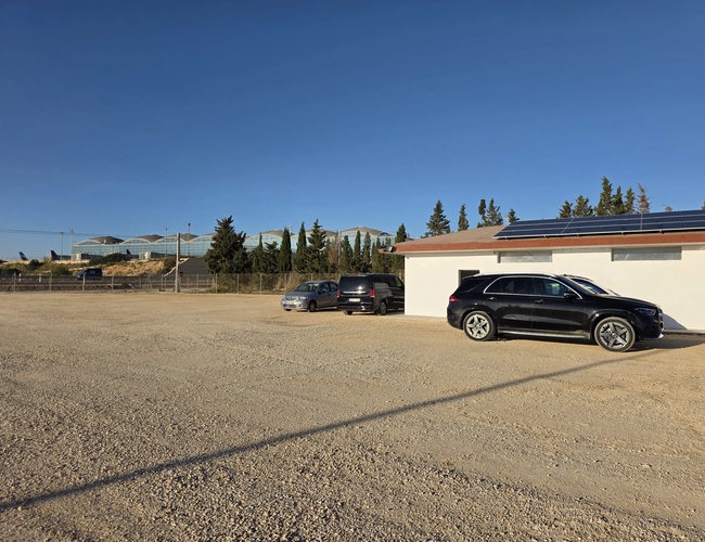 Duży, pusty, żwirowy parking z jednym zaparkowanym czarnym samochodem, w tle budynki i drzewa, pod jasnym, niebieskim niebem.