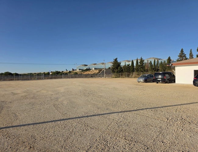 Pusty żwirowy plac z kilkoma samochodami przy budynku, widok z podobnej perspektywy co zdjęcie 1, w tle drzewa i niebieskie niebo.