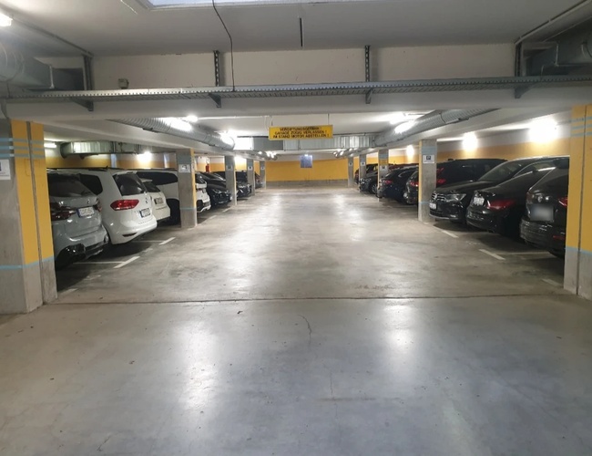 Podziemny parking z zaparkowanymi samochodami wzdłuż ściany, dobrze oświetlony przestronny korytarz.