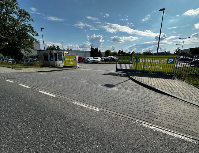 Widok na bramę wjazdową parkingu. W tle widok samochodów zaparkowanych na parkingu, którego teren jest wyłożony kostką brukową.