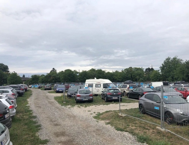 Parktiger Airport Parking 4 - Na parkingu stoją zaparkowane auta. Teren jest nieutwardzony i przestronny. W tle znajdują się drzewa.