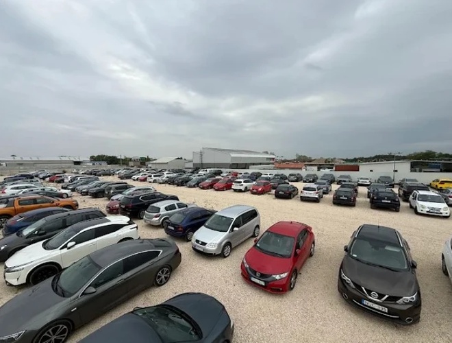 Duży parking wypełniony zaparkowanymi samochodami osobowymi na żwirowym placu, widok z lekkiej wysokości.