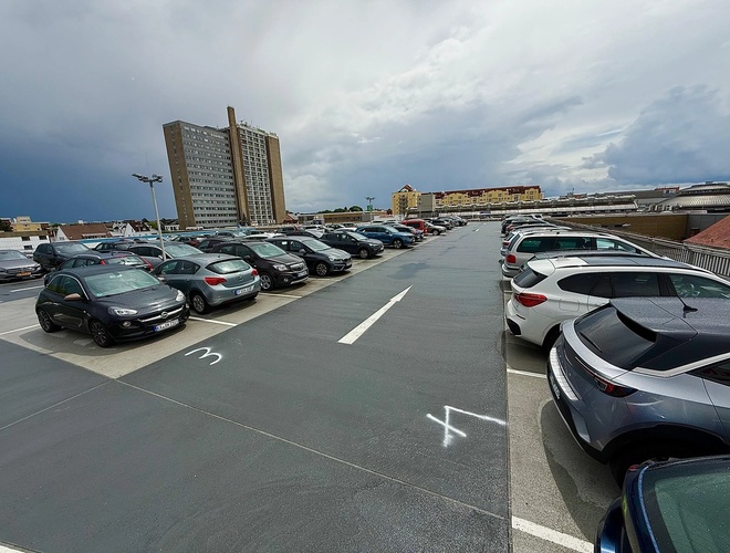 Samochody ustawione w rzędach na parkingu zewnętrznym. Asfaltowa powierzchnia, dookoła budynki.