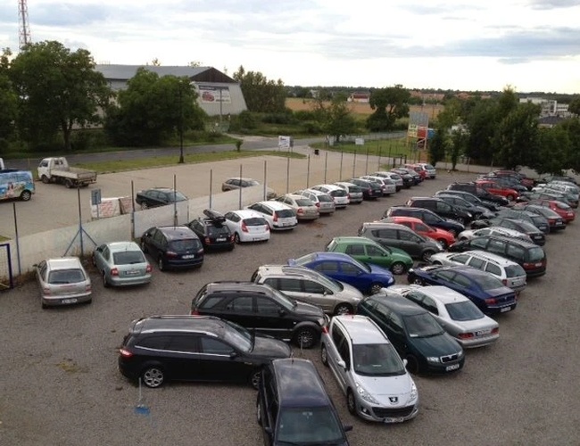 Widok z lotu ptaka na parking z wieloma zaparkowanymi samochodami w rzędach, w tle widać drzewa i budynki.