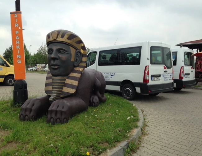 Rzeźba Sfinksa ustawiona na trawniku przy wjeździe na parking, w tle widać biały bus i część budynku