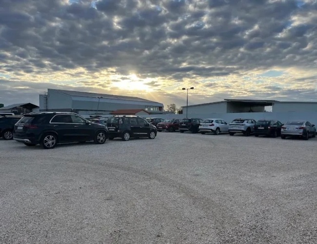 Żwirowy parking z rozproszonymi samochodami w świetle zachodzącego słońca i częściowo zachmurzonym niebem.