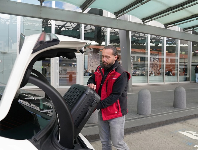 Parking Mein Valet – osoba w czerwonej kurtce ładuje bagaż do samochodu.  W tle widoczny jest teren lotniska.