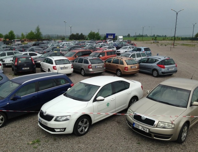 Zbliżenie na część parkingu z wieloma samochodami, w tym białym sedanem na pierwszym planie, stojącymi na utwardzonej nawierzchni.
