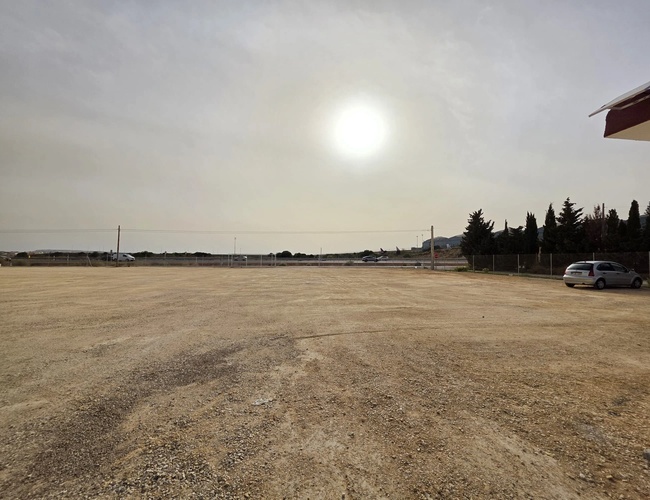 Pusty żwirowy plac pod zachodzące słońce, na horyzoncie widać drzewa i kilka oddalonych pojazdów.