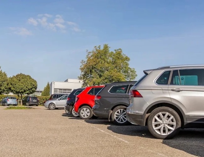 Parking Letim Brno II – na zdjęciu kilka zaparkowanych samochodów. Teren jest otoczony drzewami i budynkiem. Pogodny, słoneczny dzień.