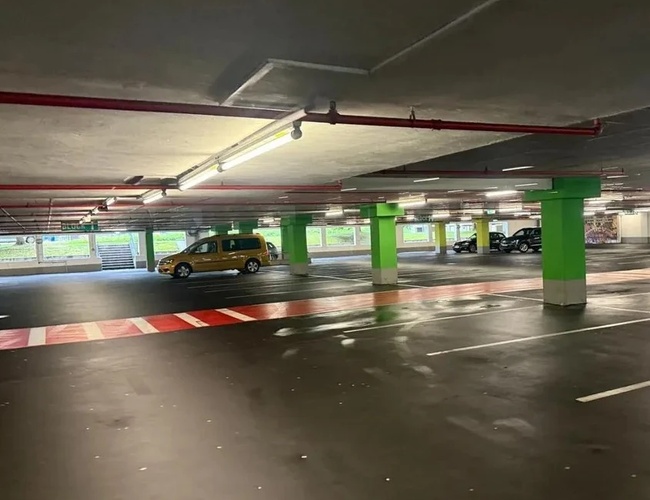 Podziemny parking z niebieskimi i zielonymi filarami, pustymi miejscami parkingowymi i kilkoma zaparkowanymi samochodami w tle.