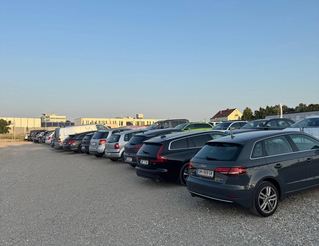 Rząd samochodów zaparkowanych na parkingu. Widoczna utwardzona nawierzchnia i budynki w tle.