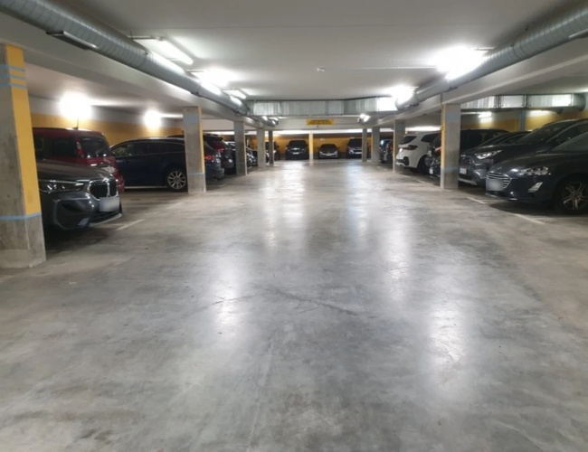 Podziemny parking z zaparkowanymi samochodami wzdłuż ściany, dobrze oświetlony przestronny korytarz.