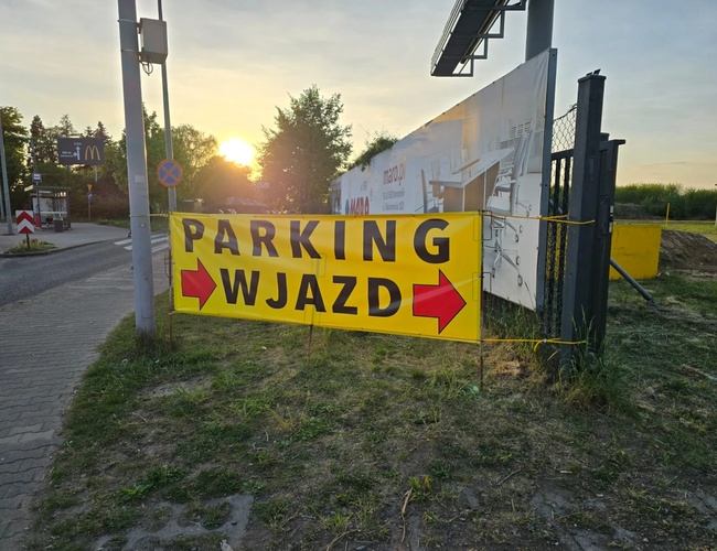 Widok wjazdu na parkingu oznaczony banerem. W oddali po lewej stronie widoczny przystanek autobusowy.