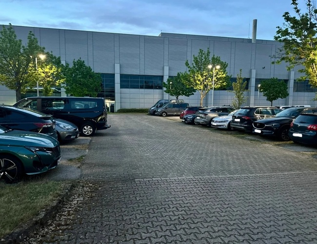 Auta zaparkowane na parkingu z kostki brukowej, dookoła drzewa i latarnie
