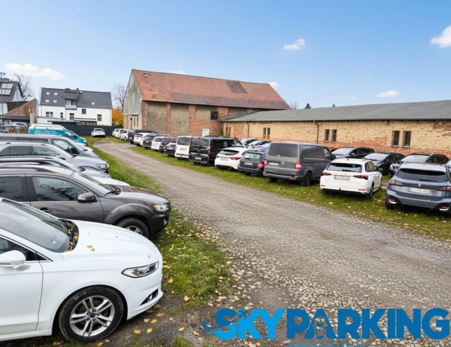 Parking na otwartej przestrzeni, położony obok dużego budynku o ceglanej fasadzie, z licznymi samochodami rozmieszczonymi wzdłuż trawiastego pasa.