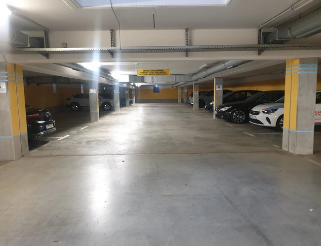 Rząd zaparkowanych samochodów w podziemnym parkingu, widoczne słupy i sufit z instalacjami.