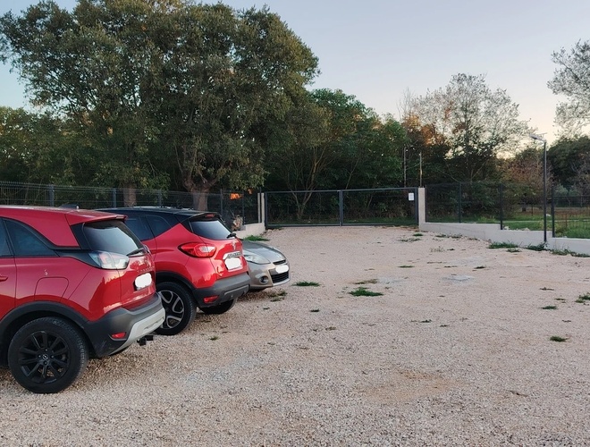Samochody zaparkowane na parkingu ze żwirowym podłożem. W tle widoczne ogrodzenie oraz drzewa. Na końcu parkingu brama wjazdowa.