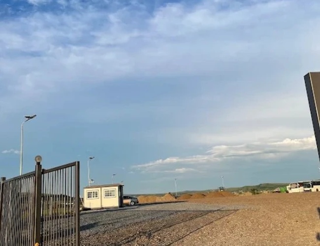 Wjazd na ogrodzony plac z kontenerem biurowym w tle i szerokim niebem z wysokimi chmurami.