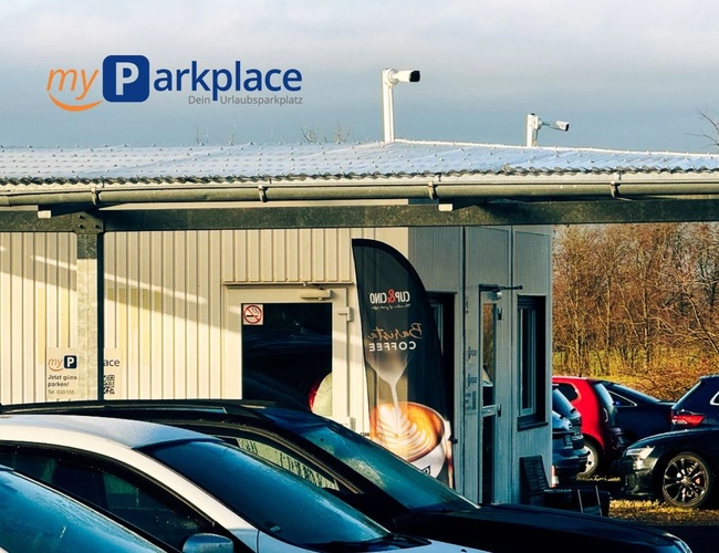Parkovisko myParkplace - Vidno niekoľko zaparkovaných áut pri budove. Na budove sú nainštalované kamery a je viditeľný reklamný plagát kávy.