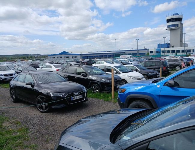 Parking P12 - Na terenie stoją liczne samochody. W tle widać budynek i wieżę kontrolną.