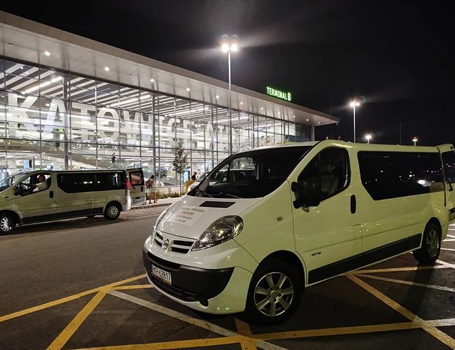 Parking con vista - Delante de la terminal hay dos furgonetas blancas. El edificio del aeropuerto está iluminado por la noche.