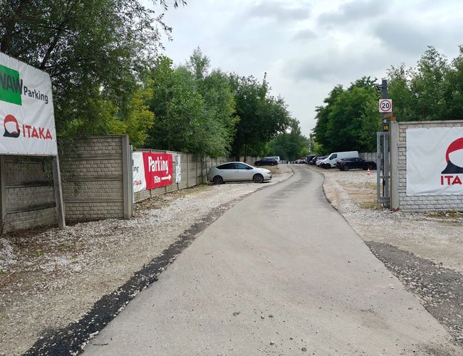 WAW Parking – Żwirowany teren z wiatą i kontenerem. Obok rosną drzewa i stoją samochody. Pogoda jest słoneczna.