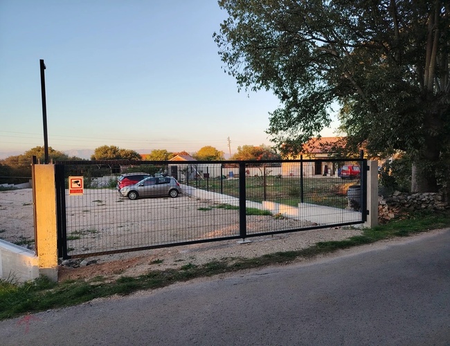 Zdjęcie bramy wjazdowej na parking. W tle kilka zaparkowanych pojazdów. Na bramie z metalową siatką znak informujący, że teren parkingu jest monitorowany.