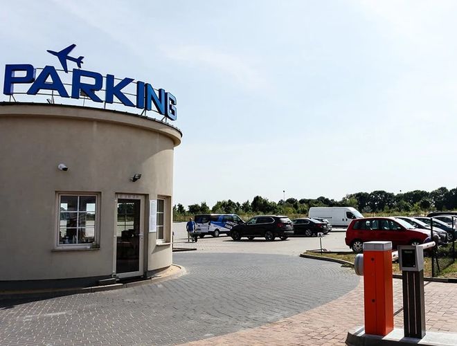 Prémium olcsó és biztonságos parkolás - Az középen áll egy épület felirattal. Mellette parkolnak autók. A terület kerítéssel körülvett és tágas.