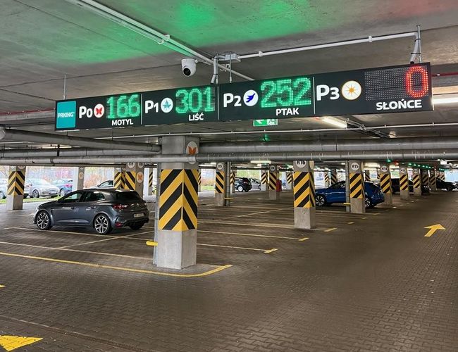 Parking P2 - Okęcie Północ Wielopoziomowy - Parking podziemny, oznaczenia z wolnymi miejscami. Kilka samochodów zaparkowanych, dużo wolnej przestrzeni.