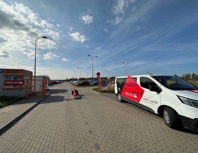 Odkryj-Parking GDN - Samochód stoi obok ogrodzonego terenu. Parking jest wyłożony kostką. Na niebie są chmury.