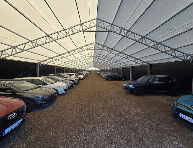 AleParking zadaszony - Wielu samochodów pod zadaszeniem. Parking jest żwirowy i ogrodzony. Konstrukcja dachu z metalowych belek.